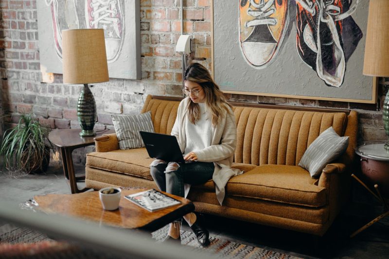 Lady wearing glasses sitting on a mustard coloured couch. She is looking at a laptop.